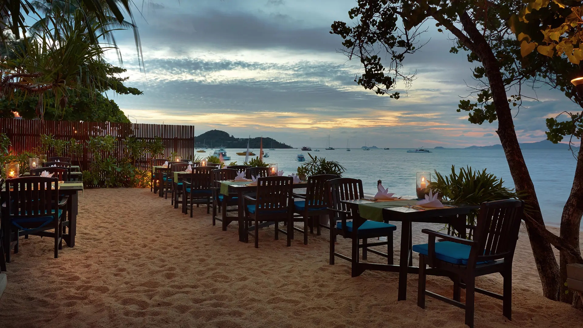 Tables set on the sand under trees, boats on the bay at sunset